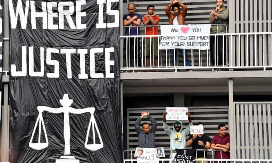 Refugees protest on a hotel balcony in Brisbane. Some hold signs and others stand with their arms crossed on their chest. There is a large black and white banner that says, "Where is Justice" that hangs beside them.