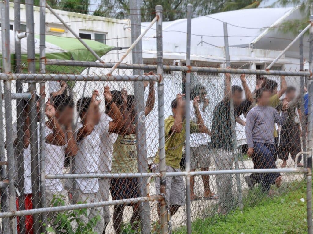 Refugees stand against a wire fence in Manus, PNG. Their faces are blurred.
